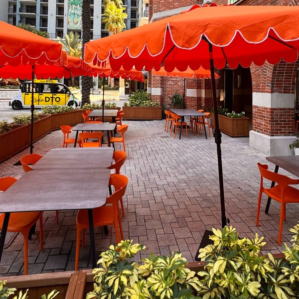 Outdoor patio of Cucina Pizza & Bar with orange umbrellas, tables, and chairs on a brick pavement.