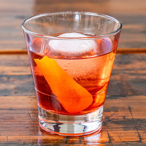 A vibrant orange cocktail with a large ice cube and an orange peel garnish on a wooden table.