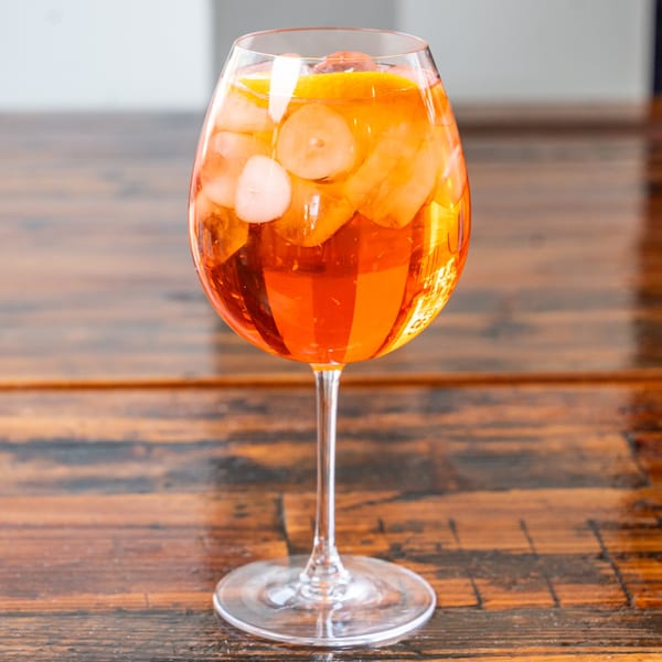 A vibrant orange Aperol Spritz cocktail with ice and orange slices in a stemmed glass on a wooden table.
