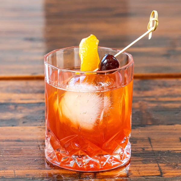 A vibrant orange cocktail with a large ice sphere, garnished with an orange peel and a cherry.