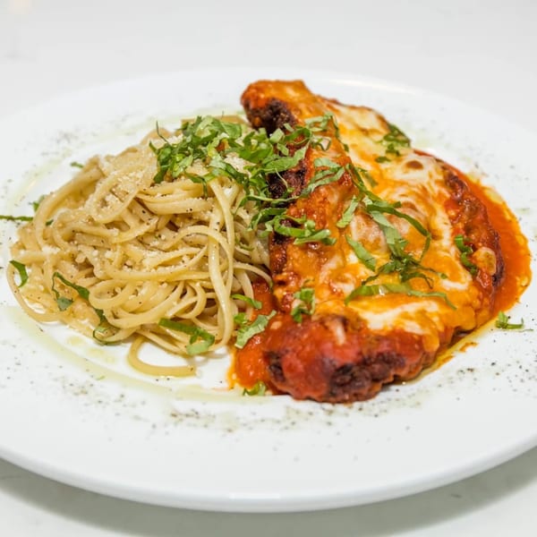 Chicken parmesan with melted cheese and red sauce, served alongside linguine pasta, garnished with fresh basil.
