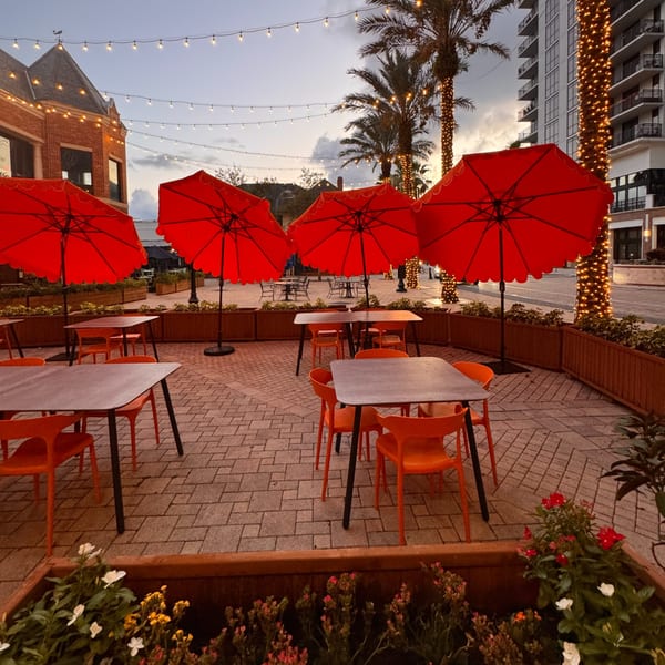 Cucina Pizza & Bar's outdoor patio with red umbrellas, tables, and string lights at dusk.