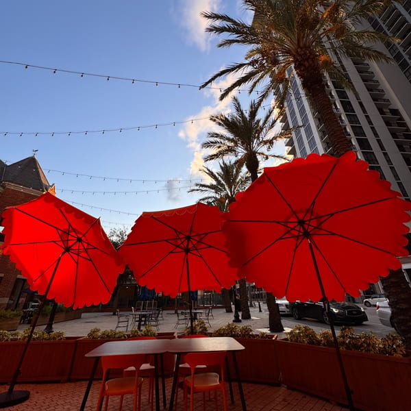 Three vibrant red umbrellas shade an outdoor dining area with string lights, palm trees, and buildings.