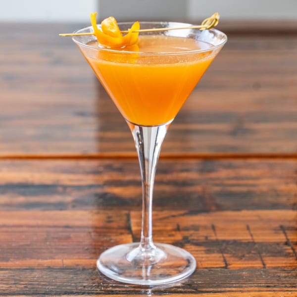 A vibrant orange cocktail in a martini glass, garnished with an orange peel, on a wooden table.