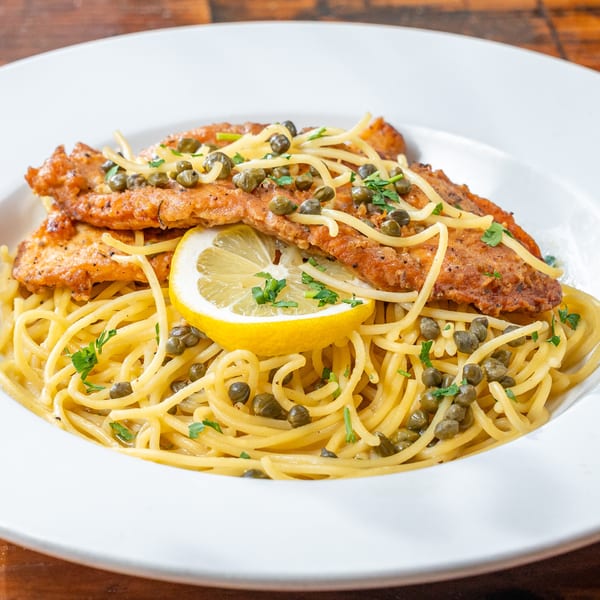 Chicken piccata with spaghetti, capers, and a lemon slice in a white bowl.