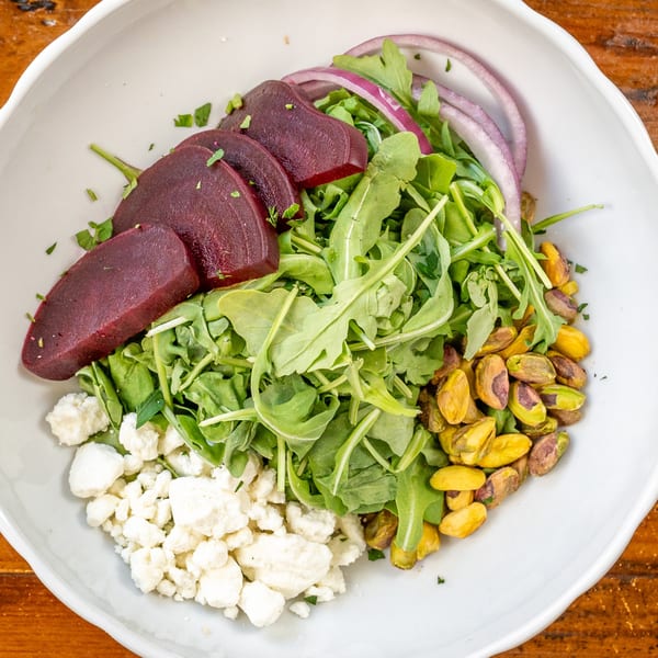 A vibrant Italian salad with sliced beets, arugula, red onion, feta cheese, and pistachios in a white bowl.