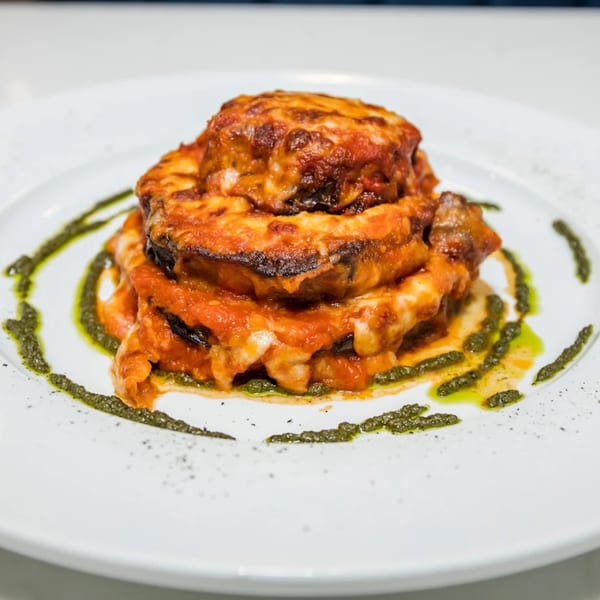 Stacked eggplant parmesan with melted cheese and red sauce, garnished with pesto and pepper on a white plate.